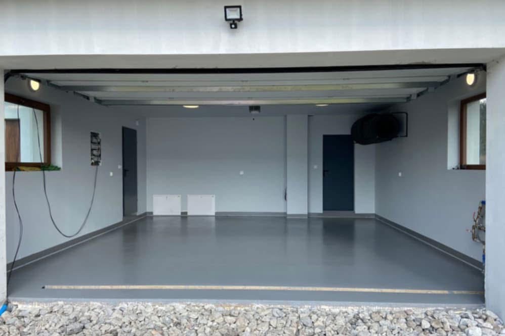 Wnętrze garażu z szarą, błyszczącą posadzką. Widoczne okna, drzwi i instalacja elektryczna na ścianie. Na dole widoczny fragment kamiennego podłoża przed garażem.