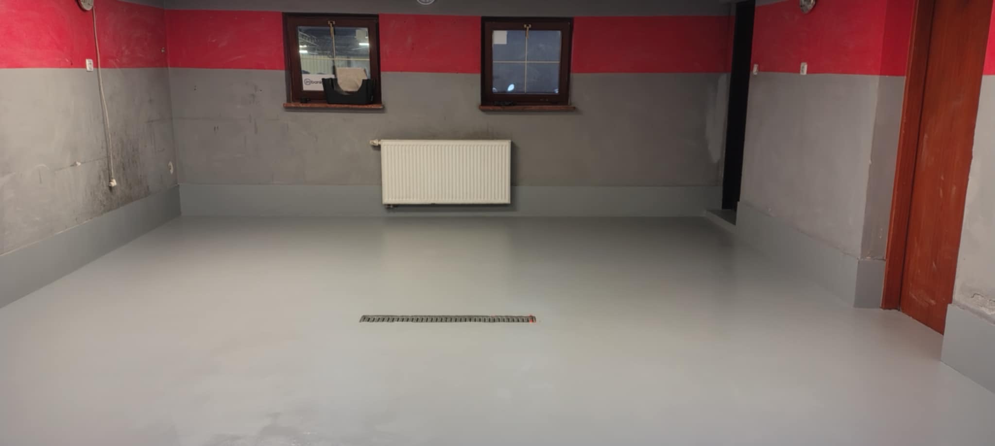 Jasnoszara, gładka powierzchnia w przemysłowym wnętrzu z grzejnikiem i odpływem liniowym w posadzce.