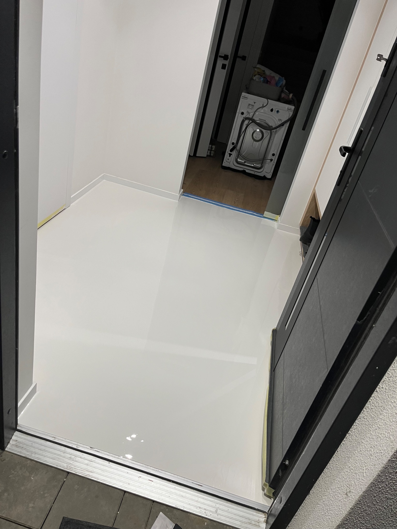 Biała, błyszcząca posadzka żywiczna w małym pomieszczeniu, widok z progu drzwi. W tle pralka i zabezpieczone taśmą malarską ściany.