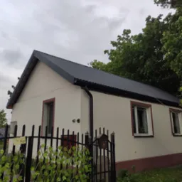 Dom jednorodzinny z nowym, grafitowym dachem z blachy trapezowej, widok zza czarnego, metalowego ogrodzenia z dekoracją z sztucznych liści.