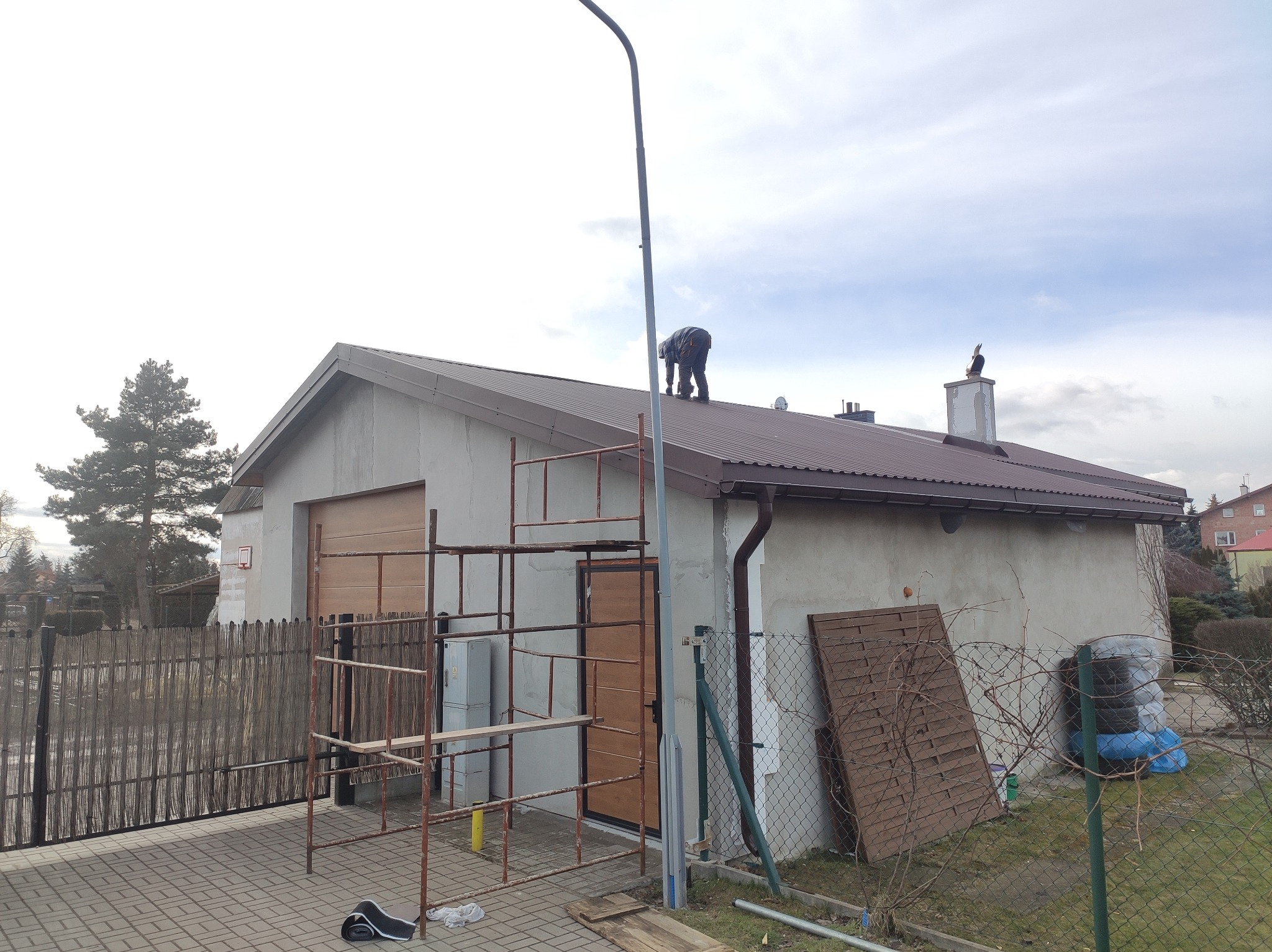 Pracownik na dachu garażu z brązową blachodachówką, podczas montażu lub naprawy, z widocznym rusztowaniem i figurką kruka na kominie.