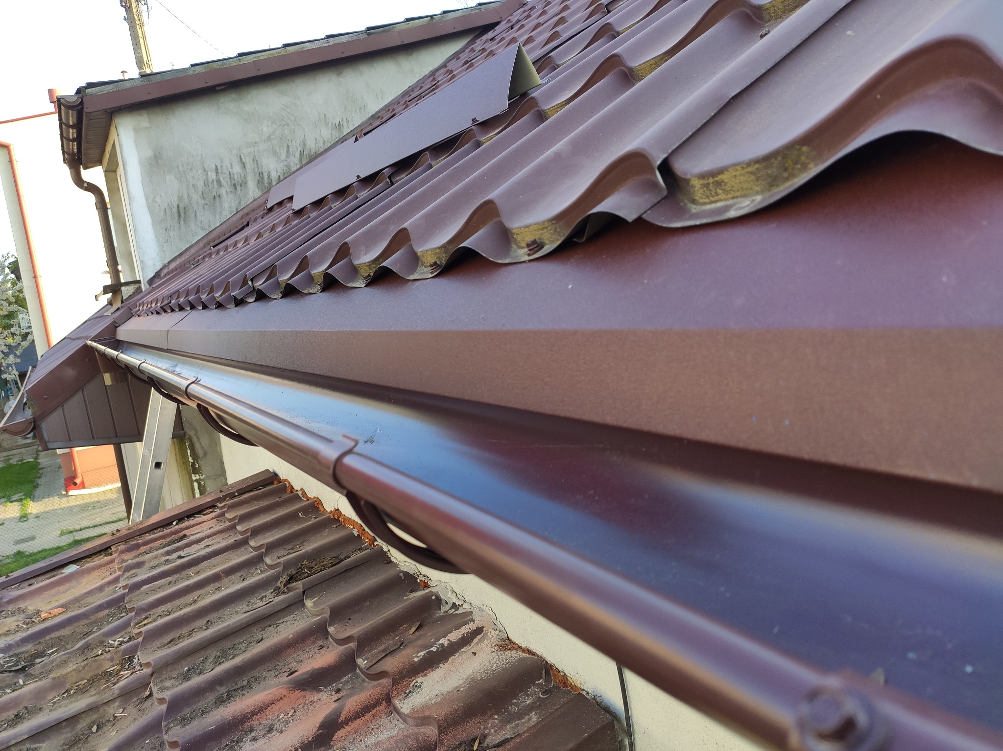 Nowe, błyszczące rynny w kolorze brązowym zamontowane na dachu z blachodachówki, widoczne elementy obróbek blacharskich i fragment starego dachu poniżej.