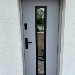 VINDOOR Damian Soja - Szare drzwi zewnętrzne z czarną, pionową szybą i klamką, osadzone w białej elewacji. Widoczny fragment chodnika i narzędzie przy podstawie ściany.