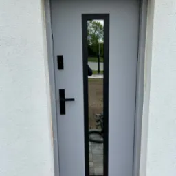 Szare drzwi zewnętrzne z czarną, pionową szybą i klamką, osadzone w białej elewacji. Widoczny fragment chodnika i narzędzie przy podstawie ściany.
