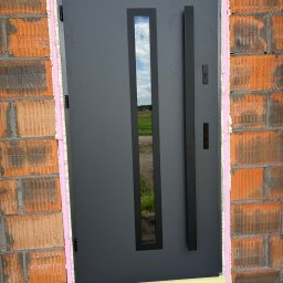 VINDOOR Damian Soja - Grafitowe drzwi zewnętrzne z długim uchwytem i wąskim przeszkleniem, osadzone w surowym murze z pianką montażową. Widok na pole przez szybę.