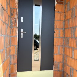 VINDOOR Damian Soja - Ciemnoszare drzwi zewnętrzne z szybą, osadzone w surowej, ceglanej wnęce. Widoczne detale montażowe i odbicie otoczenia w szybie.