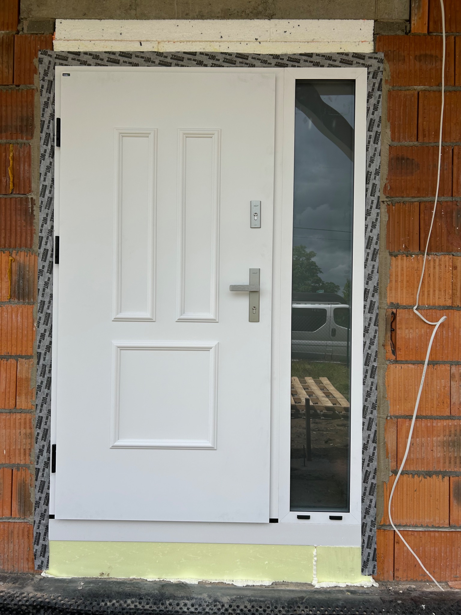 Montaż białych drzwi zewnętrznych z bocznym przeszkleniem, widoczne materiały izolacyjne i fragment ściany z cegły. Drzwi z klamką i zamkiem, w trakcie osadzania.