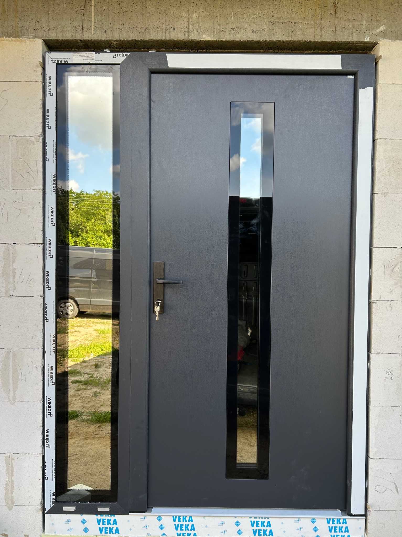 Grafitowe drzwi zewnętrzne z szybą boczną, zamontowane w surowym murze. Widoczne uszczelki i folie montażowe. Klucz w zamku. W szybie odbija się samochód.