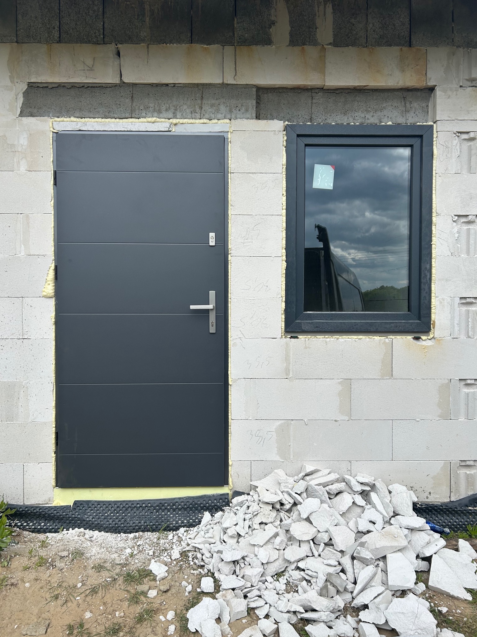 Szara brama garażowa segmentowa i okno PCV w surowym murze z betonu komórkowego w Łuczynie. Widoczna pianka montażowa i gruz budowlany.