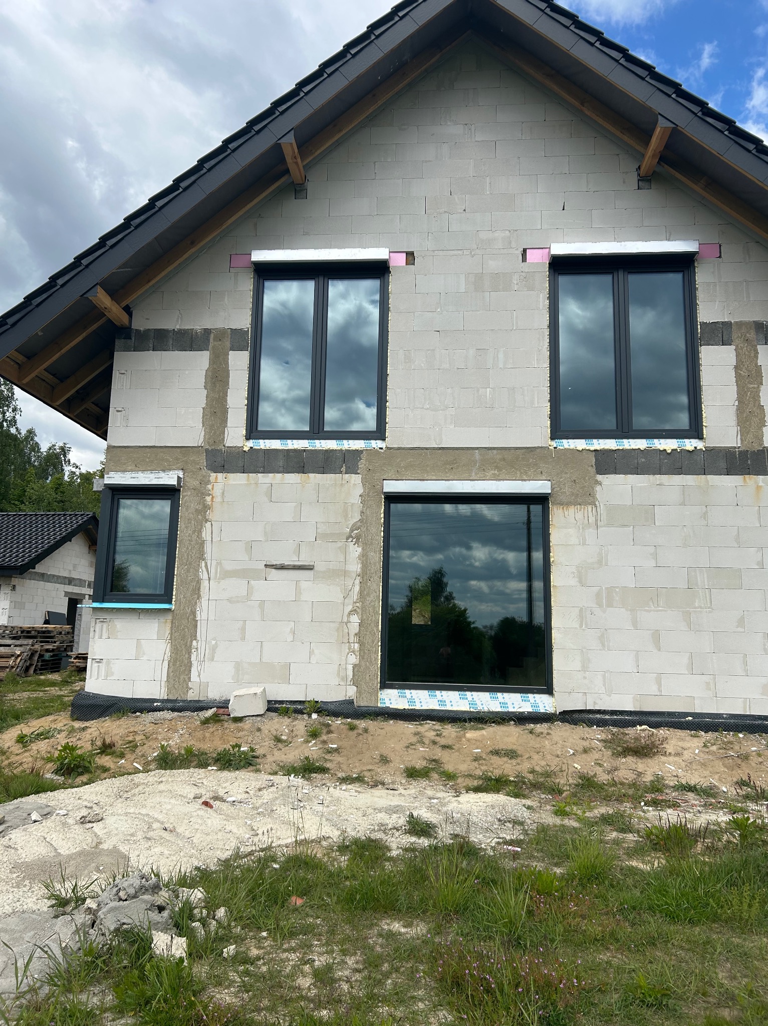 Surowy stan budynku z szarego bloczka, z zamontowanymi oknami PCV w czarnych ramach. Widoczne przygotowania do montażu rolet zewnętrznych.