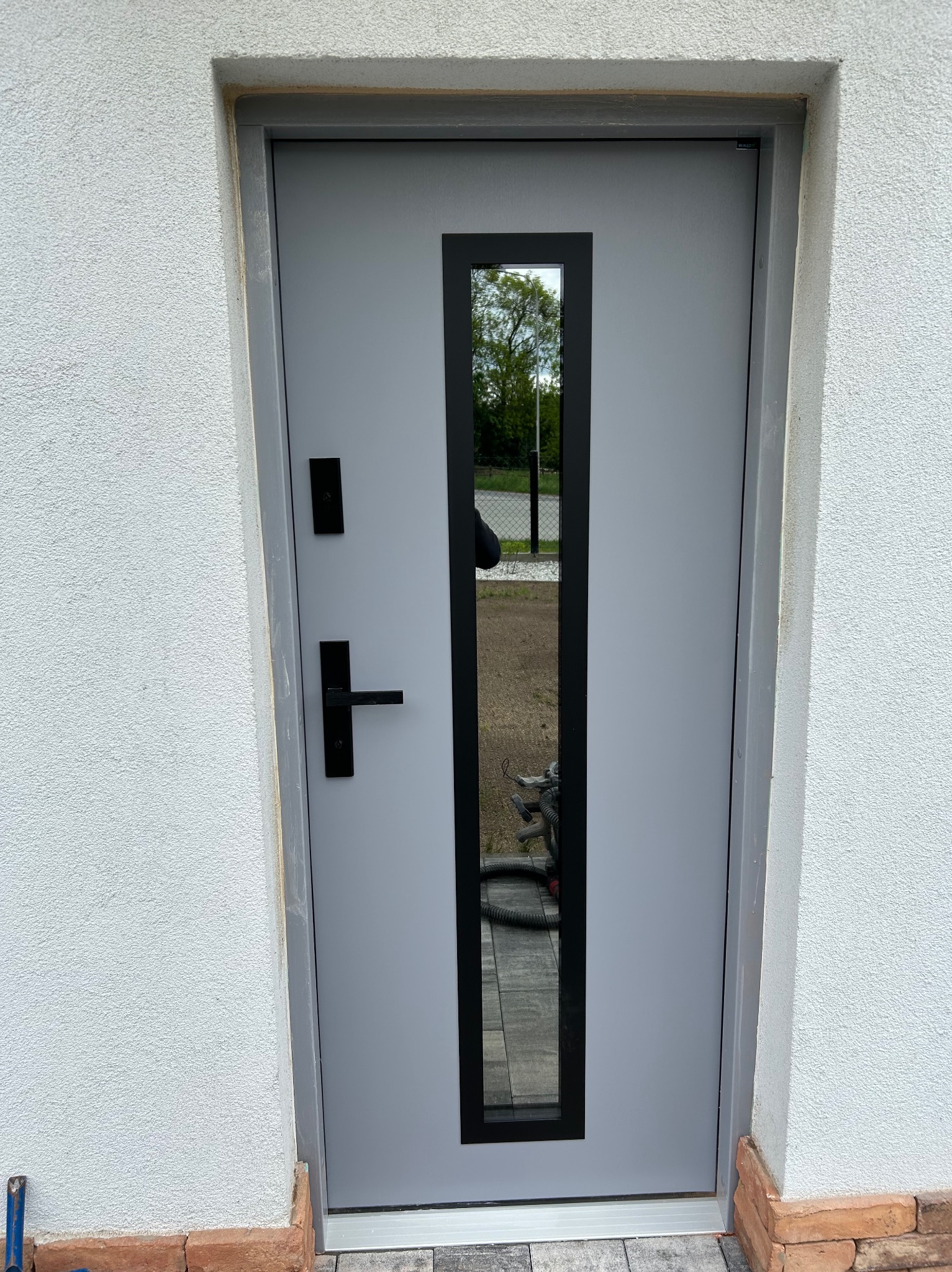 Szare drzwi zewnętrzne z czarną, pionową szybą i klamką, osadzone w białej elewacji. Widoczny fragment chodnika i narzędzie przy podstawie ściany.