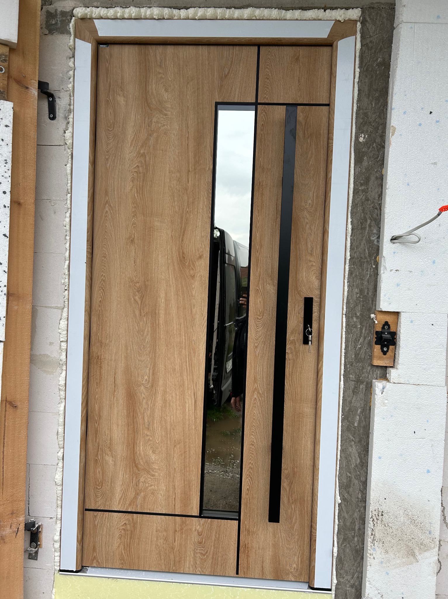 Montaż drzwi zewnętrznych z szybą i drewnopodobnym dekorem, widoczne pianka montażowa i elementy mocujące, klucz w zamku. Drzwi w trakcie osadzania w surowym otworze.