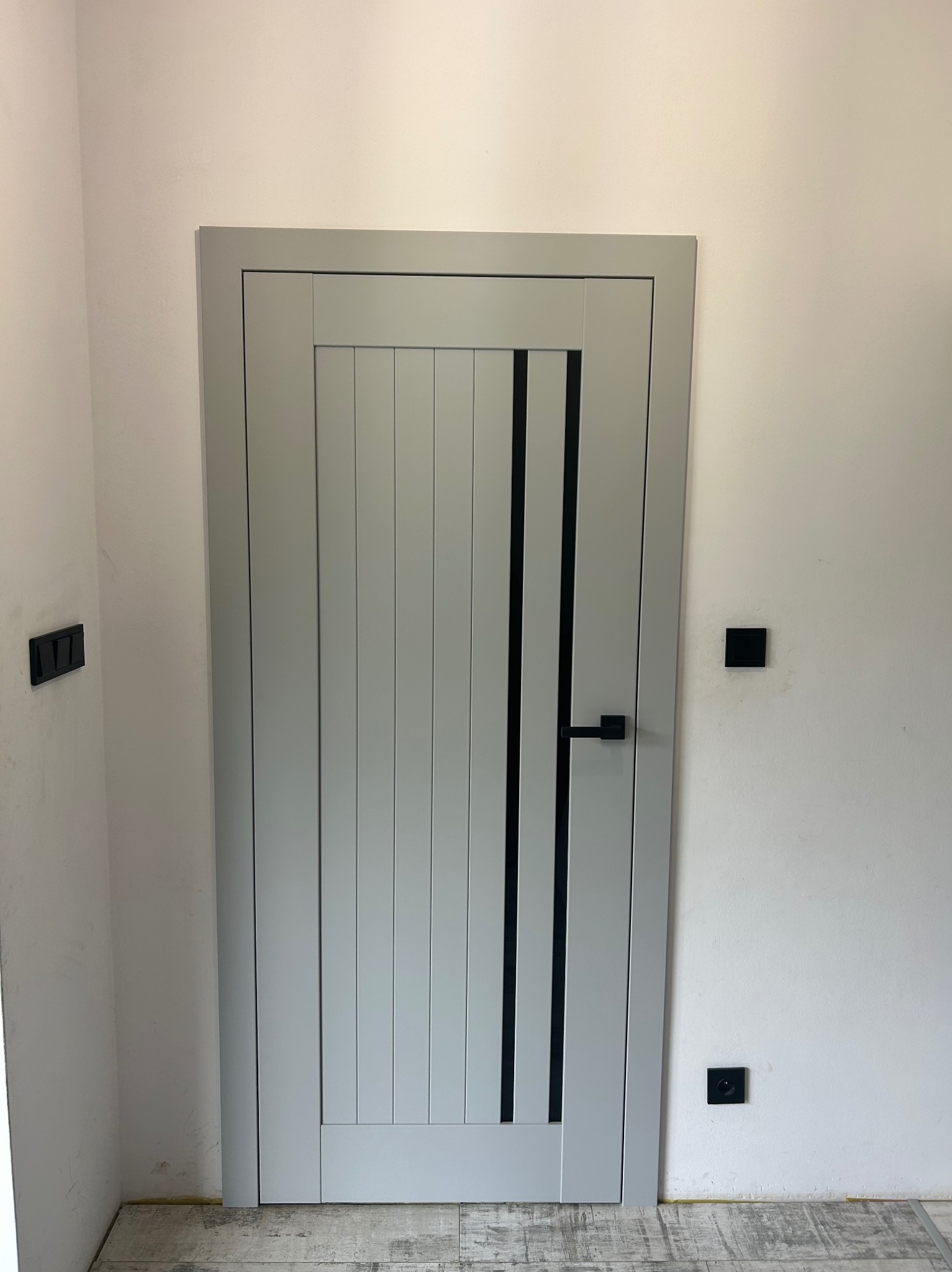Szare drzwi wewnętrzne z pionowymi panelami i czarnymi akcentami, czarna klamka, widoczne czarne włączniki na ścianie, podłoga w odcieniach szarości i beżu.