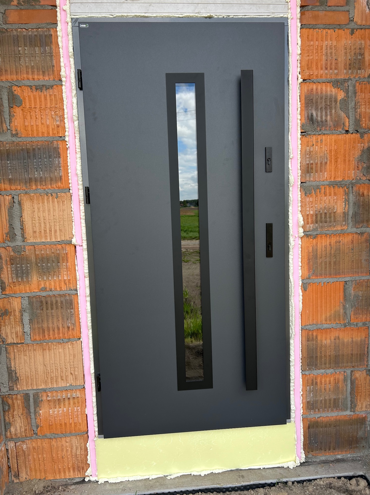 Grafitowe drzwi zewnętrzne z długim uchwytem i wąskim przeszkleniem, osadzone w surowym murze z pianką montażową. Widok na pole przez szybę.