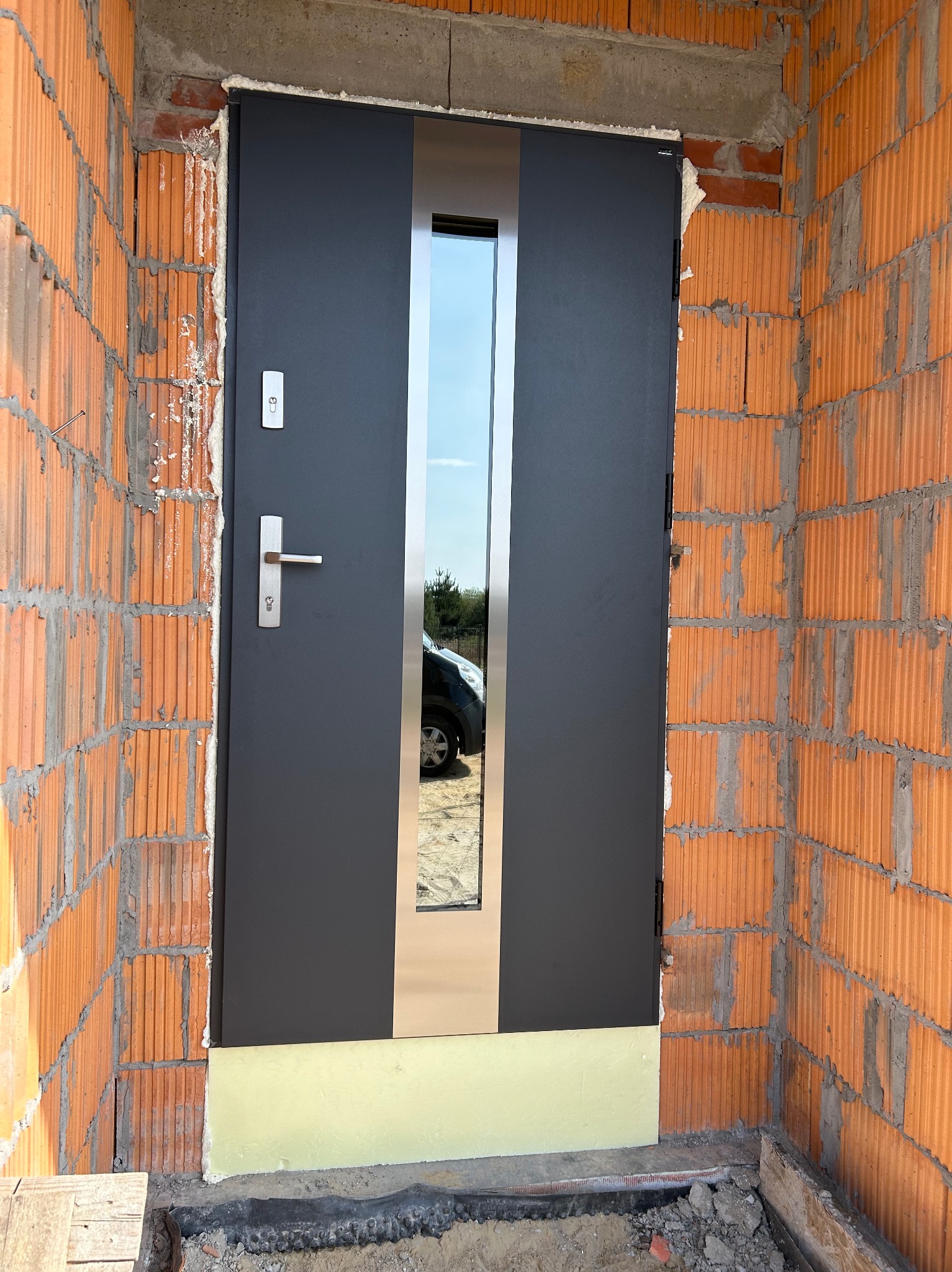 Ciemnoszare drzwi zewnętrzne z szybą, osadzone w surowej, ceglanej wnęce. Widoczne detale montażowe i odbicie otoczenia w szybie.