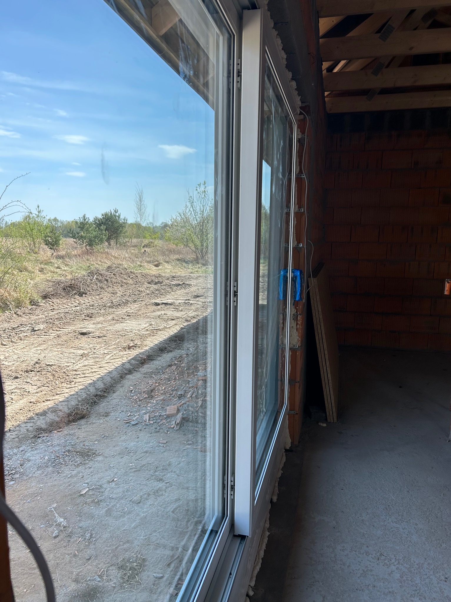 Nowe okno PCV z widokiem na zewnątrz w surowym stanie budynku. Widoczne cegły i instalacje. Okno przesuwne z białymi ramami, z zewnątrz widok na pole i drzewa.