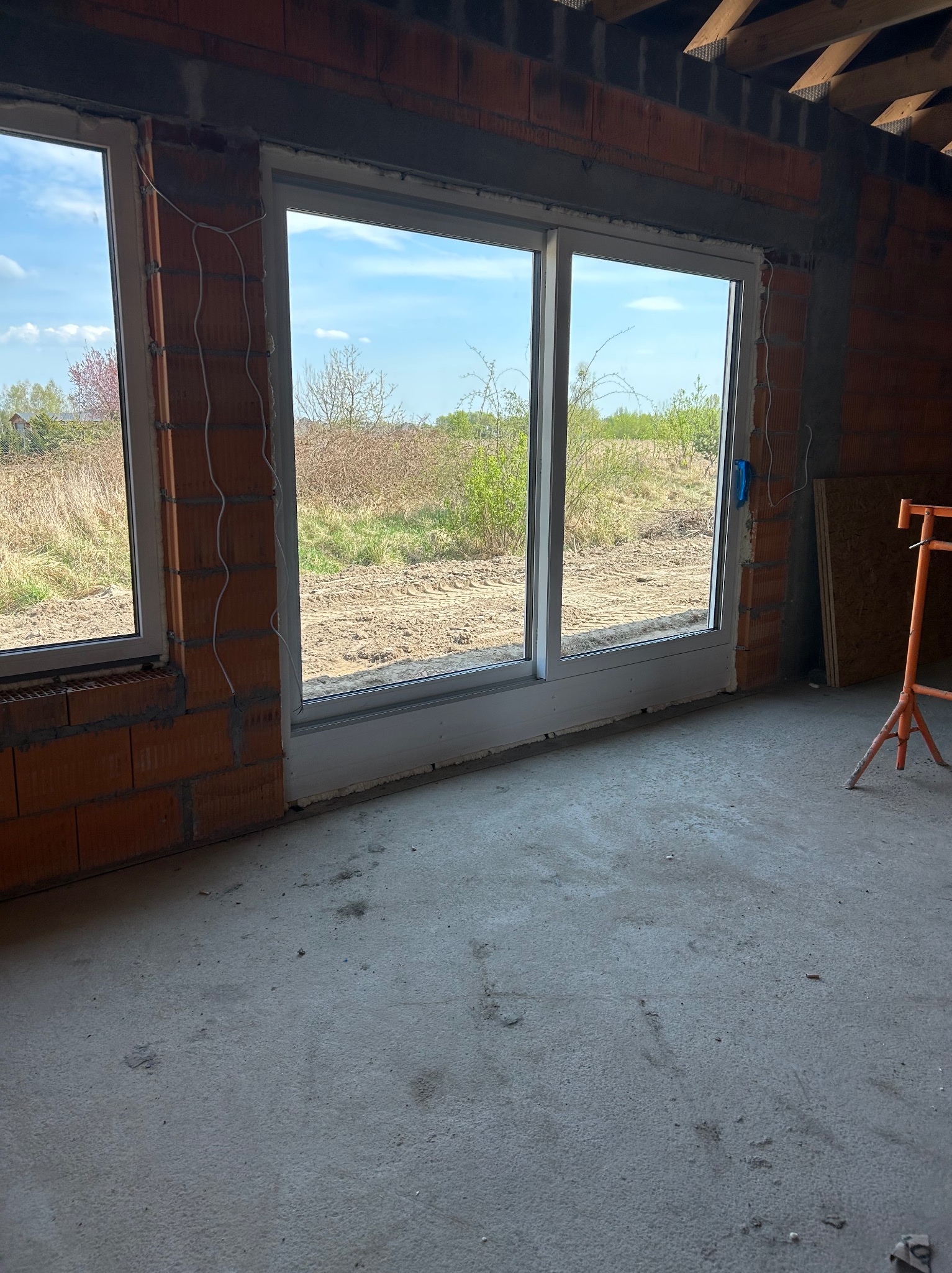 Wnętrze w trakcie budowy z widokiem na krajobraz przez białe okno. Widoczne cegły, przewody i surowe wykończenie podłogi.