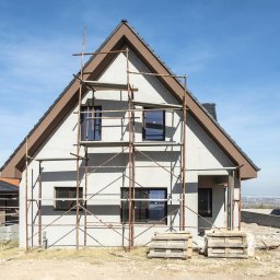 Budowa domów, remonty, usługi budowlane, ocieplanie - Dom jednorodzinny w trakcie ocieplania, widoczne rusztowania, surowy tynk, brązowe obramowania dachu i okien, palety na ziemi, w tle zabudowa miejska.