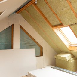 Budowa domów, remonty, usługi budowlane, ocieplanie - Poddasze w trakcie ocieplania: widoczna wełna mineralna między krokwiami, płyty gipsowo-kartonowe częściowo zamontowane, okno dachowe w tle.