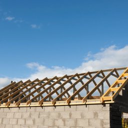 Budowa domów, remonty, usługi budowlane, ocieplanie - Surowy szkielet dachu z drewnianych belek, połączonych metalowymi płytkami, wzniesiony na ścianach z szarego betonu komórkowego, na tle błękitnego nieba z białymi obłokami.