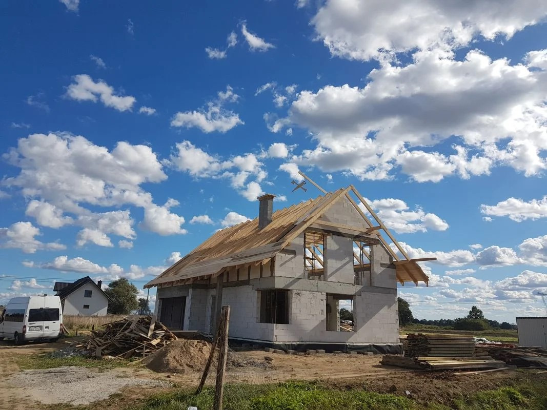 Budowa domu jednorodzinnego w trakcie: ściany z betonu komórkowego, drewniana konstrukcja dachu, widoczne składowane materiały budowlane i biały bus na tle błękitnego nieba z obłokami.