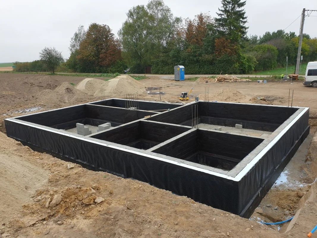 Fundamenty domu jednorodzinnego z izolacją pionową, widoczne zbrojenia, bloczki betonowe, teren budowy z hałdami piasku i przenośną toaletą w tle.