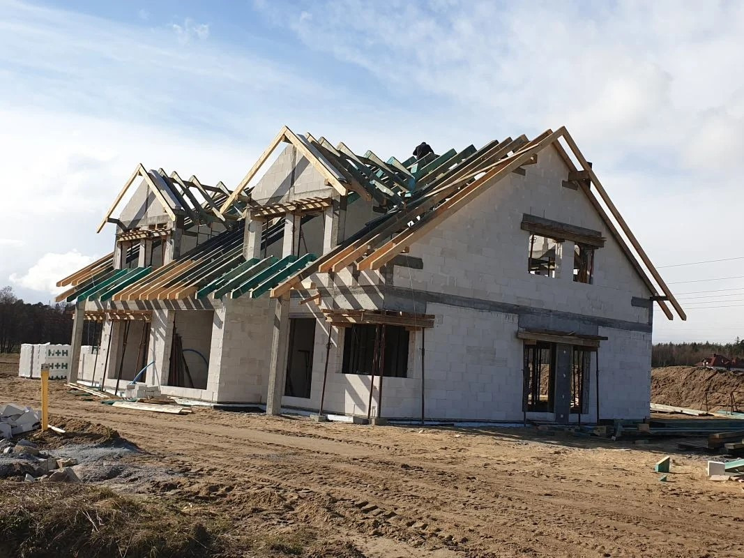 Budowa domu jednorodzinnego w trakcie realizacji: ściany z betonu komórkowego, drewniana konstrukcja dachu, widoczny pracownik na dachu, teren budowy z piaskiem.