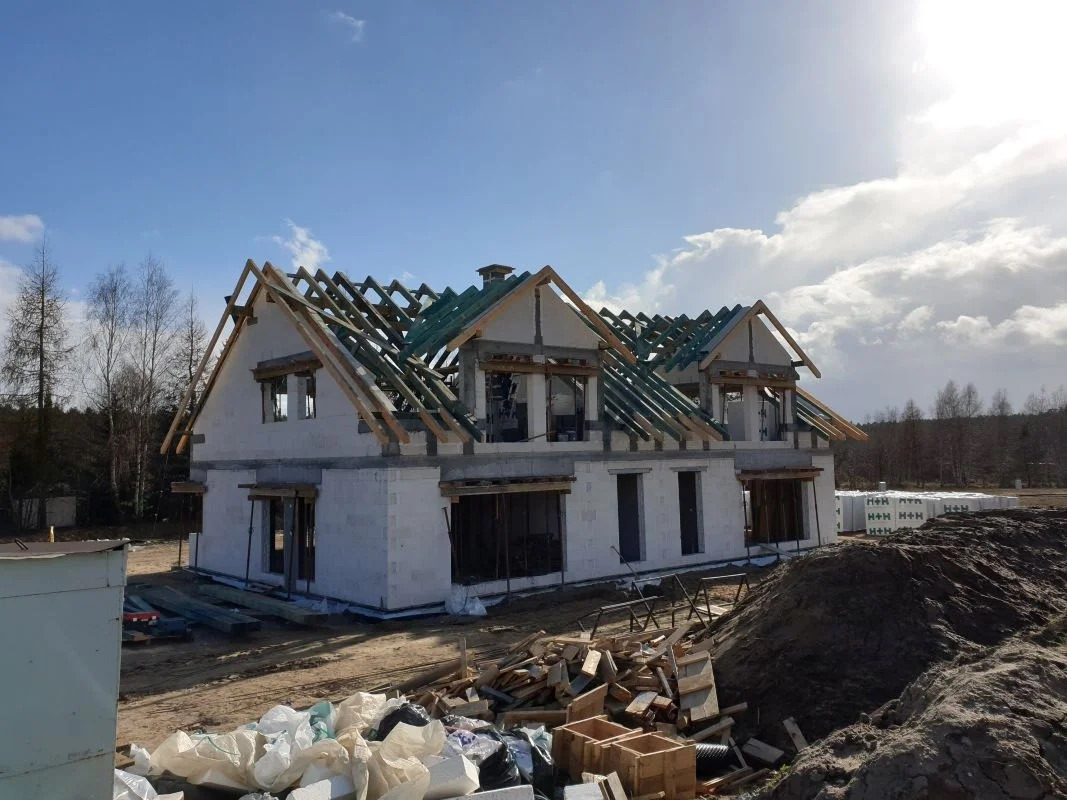 Surowy stan dwukondygnacyjnego domu z białego bloczka, z widoczną konstrukcją drewnianą dachu i zieloną membraną, na tle błękitnego nieba z chmurami i stertą materiałów budowlanych.