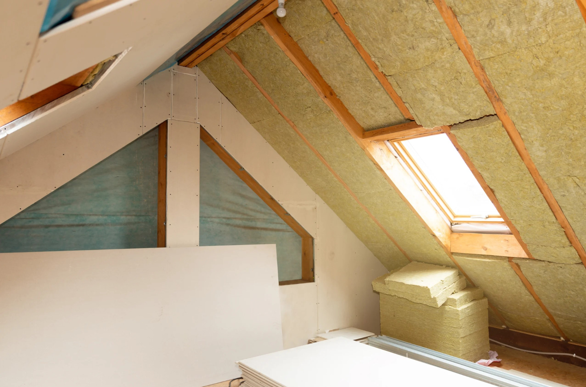 Poddasze w trakcie ocieplania: widoczna wełna mineralna między krokwiami, płyty gipsowo-kartonowe częściowo zamontowane, okno dachowe w tle.