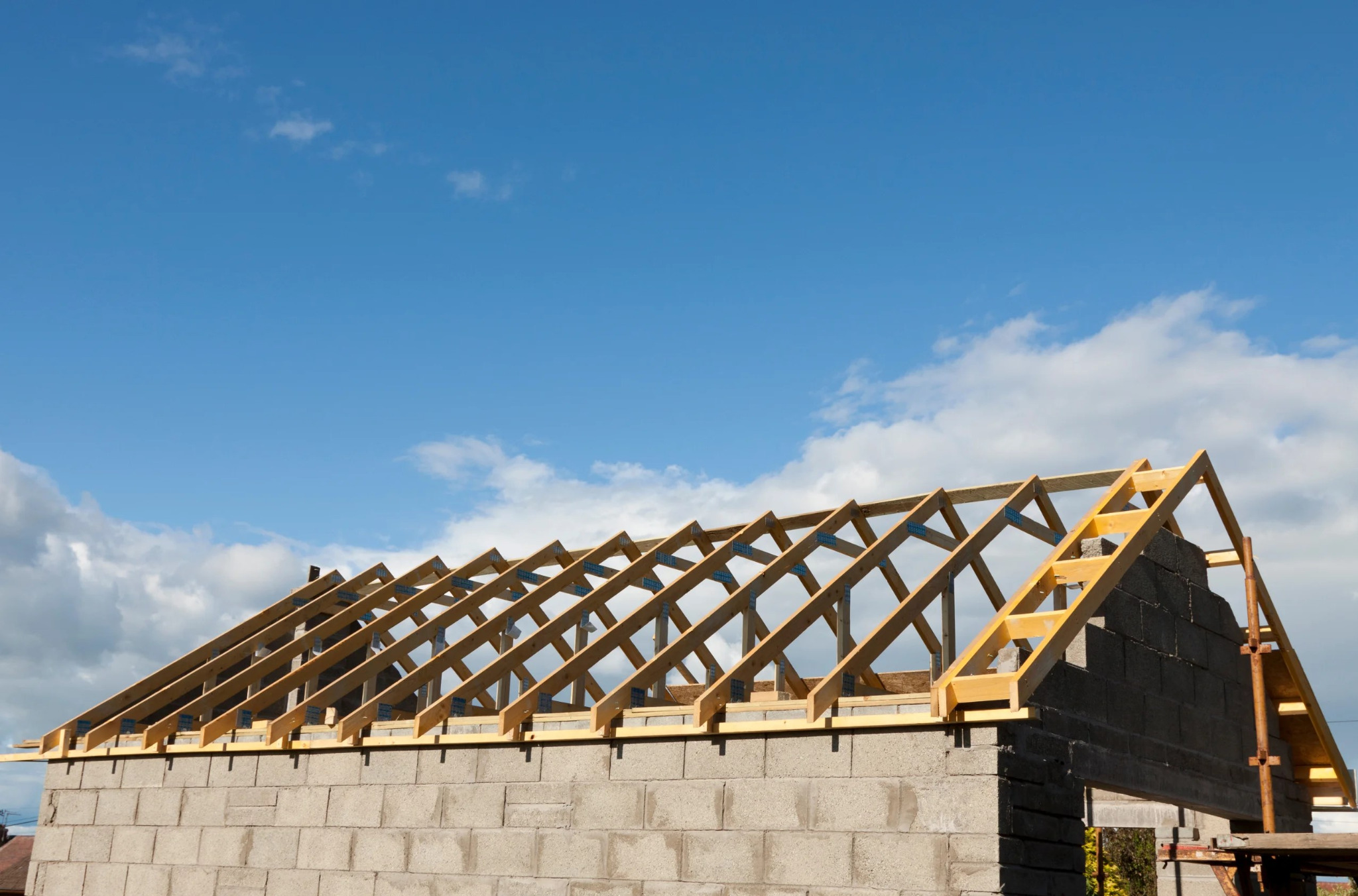 Surowy szkielet dachu z drewnianych belek, połączonych metalowymi płytkami, wzniesiony na ścianach z szarego betonu komórkowego, na tle błękitnego nieba z białymi obłokami.