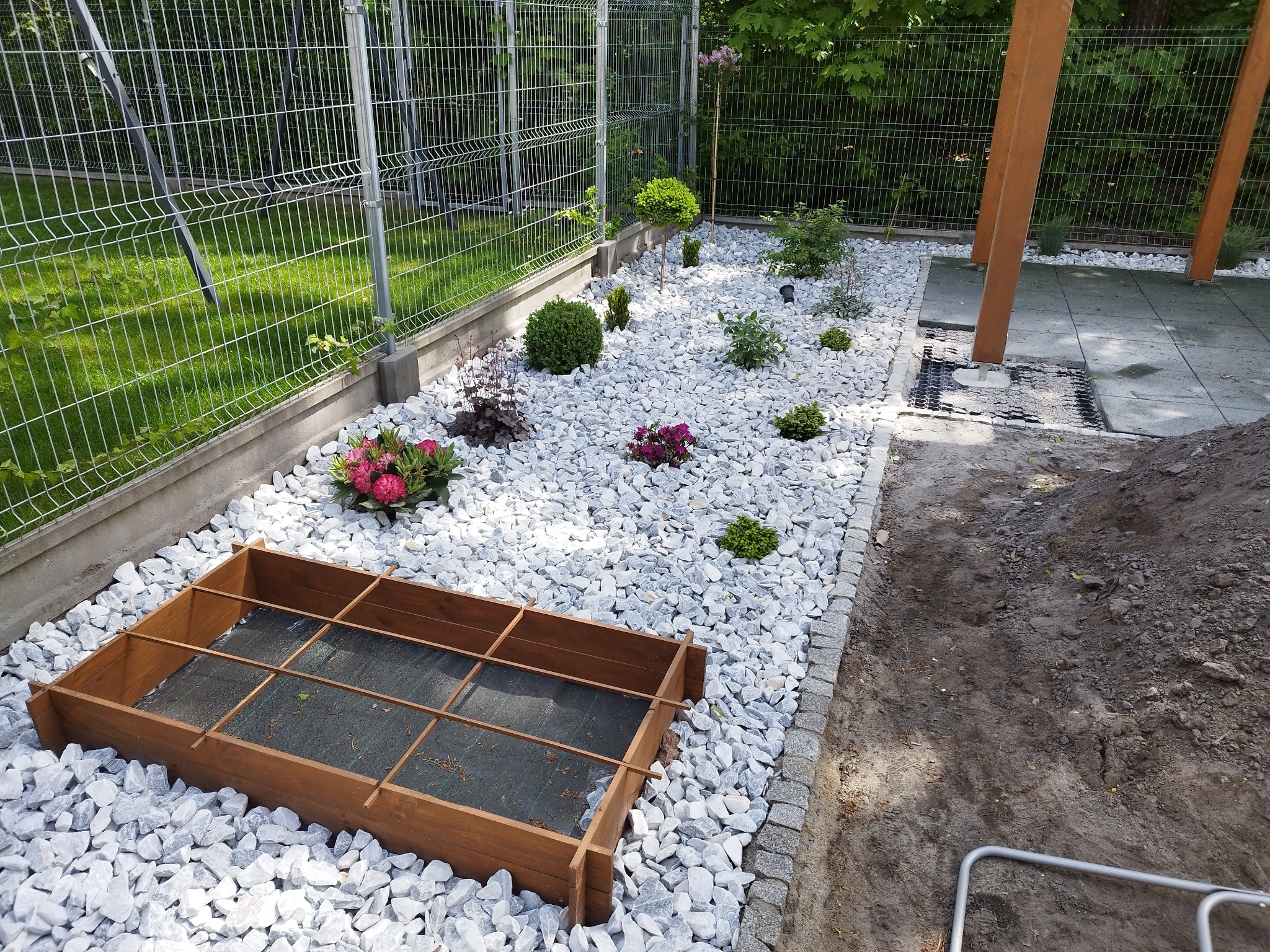Przygotowany teren pod rabatę kwiatową z drewnianą obudową, wysypany białymi kamieniami, z widocznymi nasadzeniami roślin ozdobnych i siatką ogrodzeniową w tle.