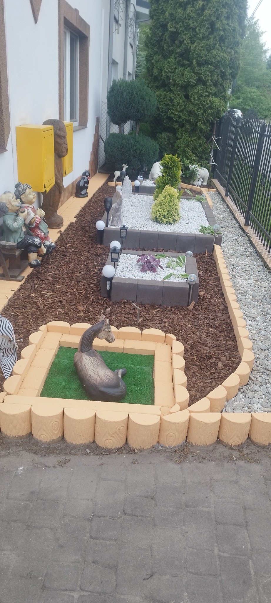 Ogród przydomowy z dekoracyjnymi rabatami wykończonymi obrzeżami z imitacji drewna, wypełnionymi korą, białym kamieniem i roślinami ozdobnymi, wzbogacony o figury ogrodowe, w tym leżącego konia.