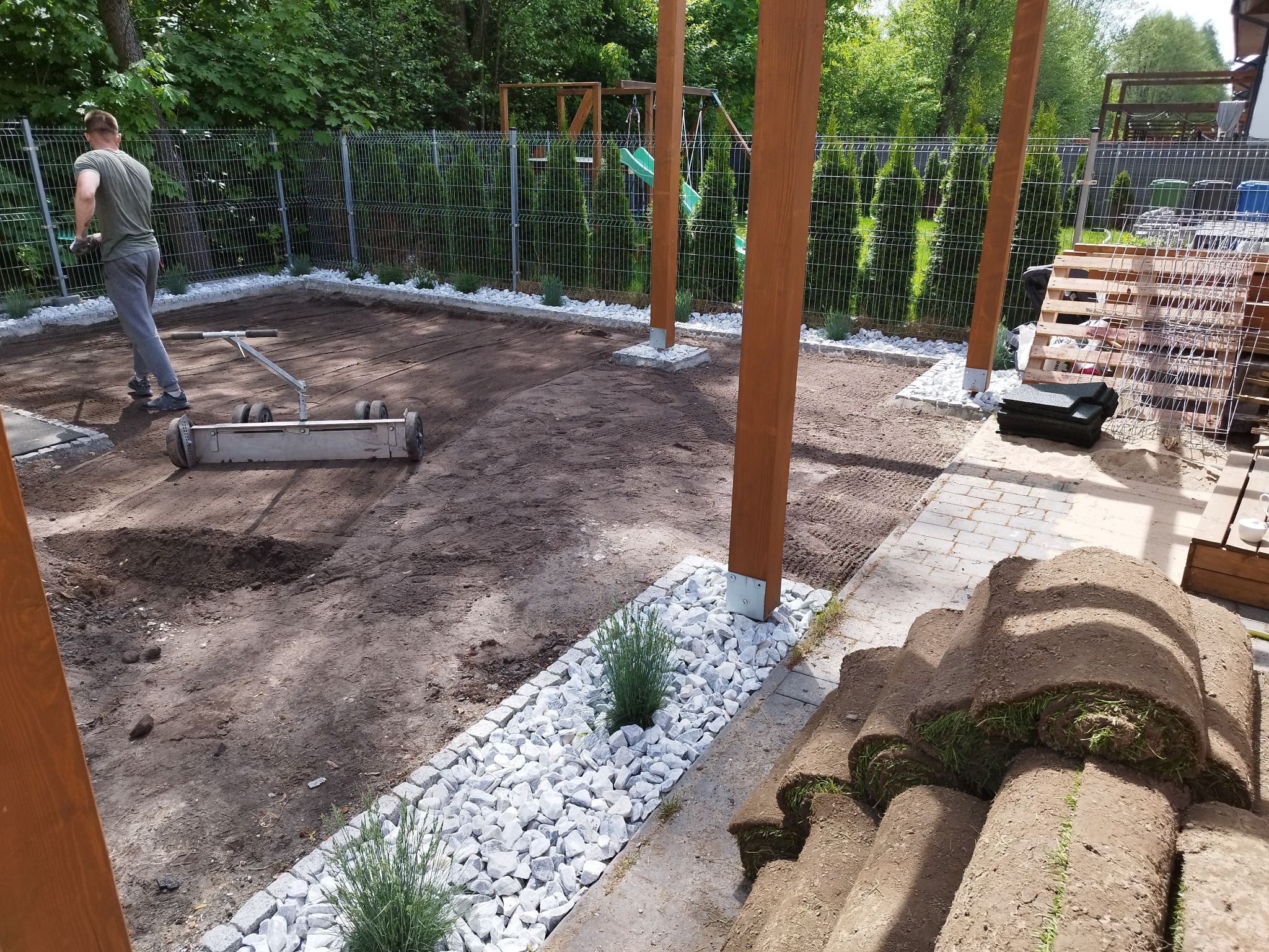 Przygotowanie terenu pod trawnik z rolki: mężczyzna wyrównuje ziemię walcem, widoczne rolki trawy, białe kamienie dekoracyjne, drewniana konstrukcja z placem zabaw w tle.