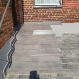 Zewnętrzne schody wyłożone szarą terakotą prowadzące do ceglanego budynku z oknem. Ciemnoszara kostka brukowa na podłożu. Widoczny cień osoby fotografującej.