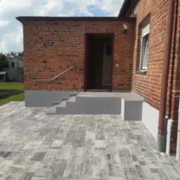 Wykończone wejście do budynku z cegły z szarymi schodami i kostką brukową. Widoczne detale, takie jak poręcz i lampa. Słoneczny dzień, błękitne niebo z chmurami.