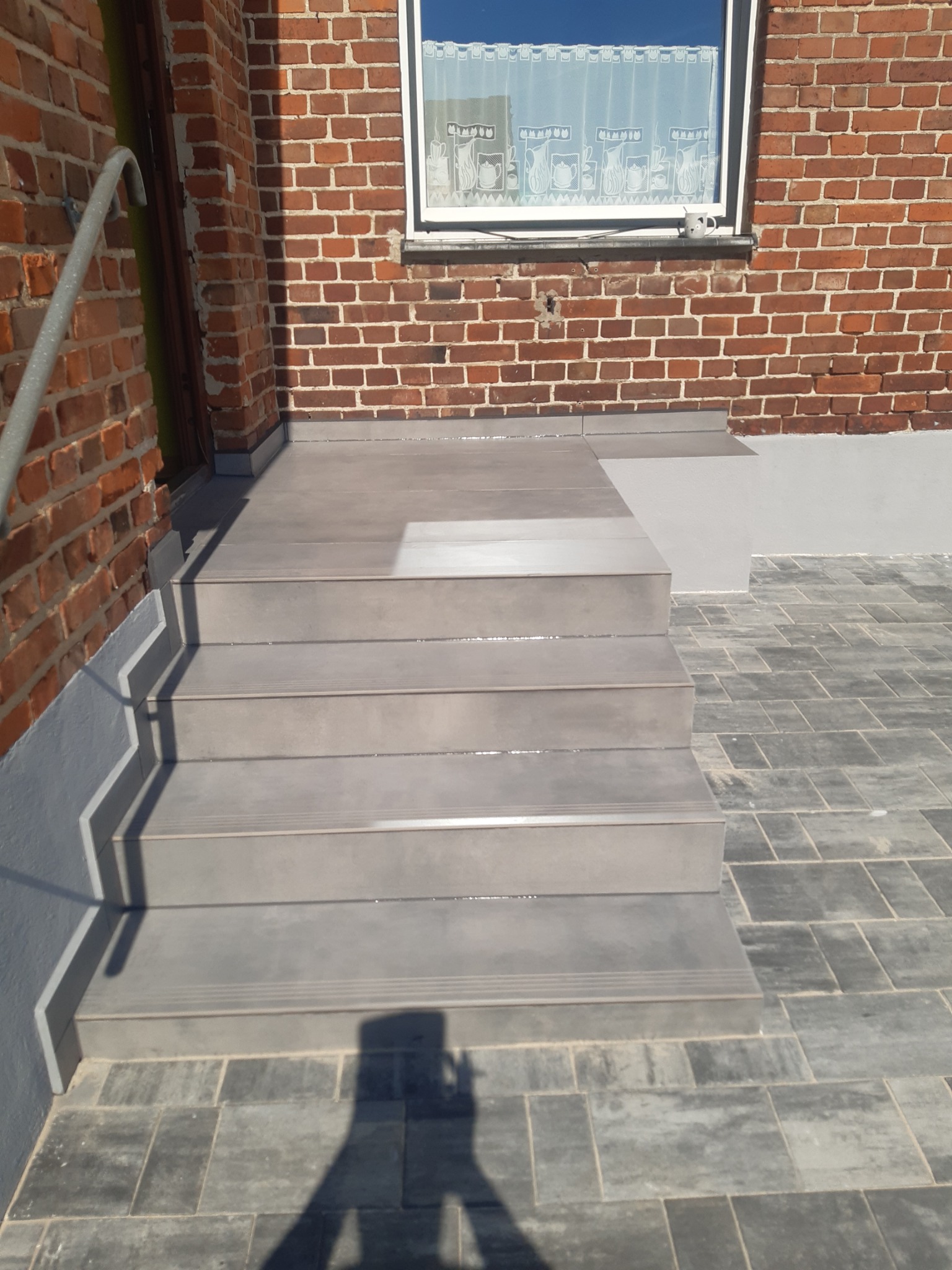 Zewnętrzne schody wyłożone szarą terakotą prowadzące do ceglanego budynku z oknem. Ciemnoszara kostka brukowa na podłożu. Widoczny cień osoby fotografującej.