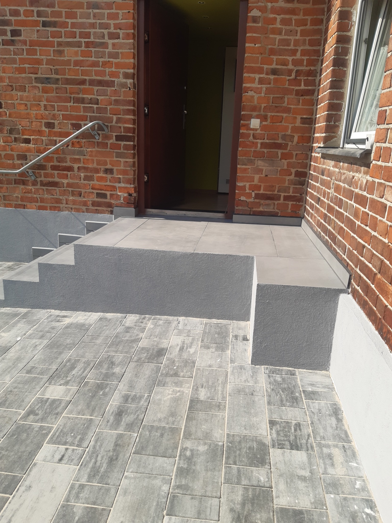 Wykończone płytkami schody wejściowe do budynku z cegły, z szarą kostką brukową na zewnątrz. Widoczne detale wykończenia i barierka ze stali nierdzewnej.