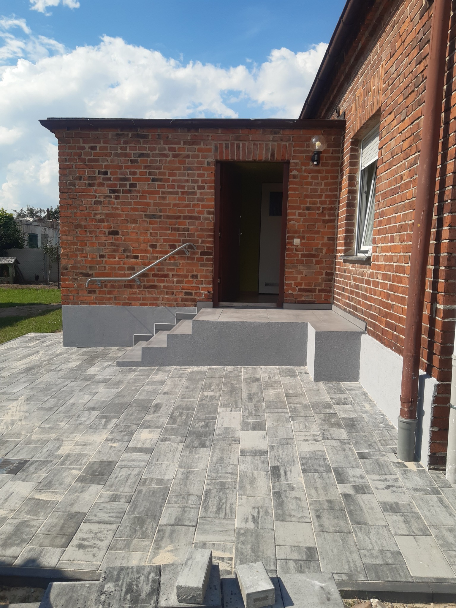Wykończone wejście do budynku z cegły z szarymi schodami i kostką brukową. Widoczne detale, takie jak poręcz i lampa. Słoneczny dzień, błękitne niebo z chmurami.