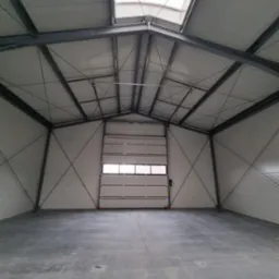 Wnętrze pustego, nowo wybudowanego magazynu z betonową posadzką, stalową konstrukcją nośną i częściowo otwartą bramą segmentową.