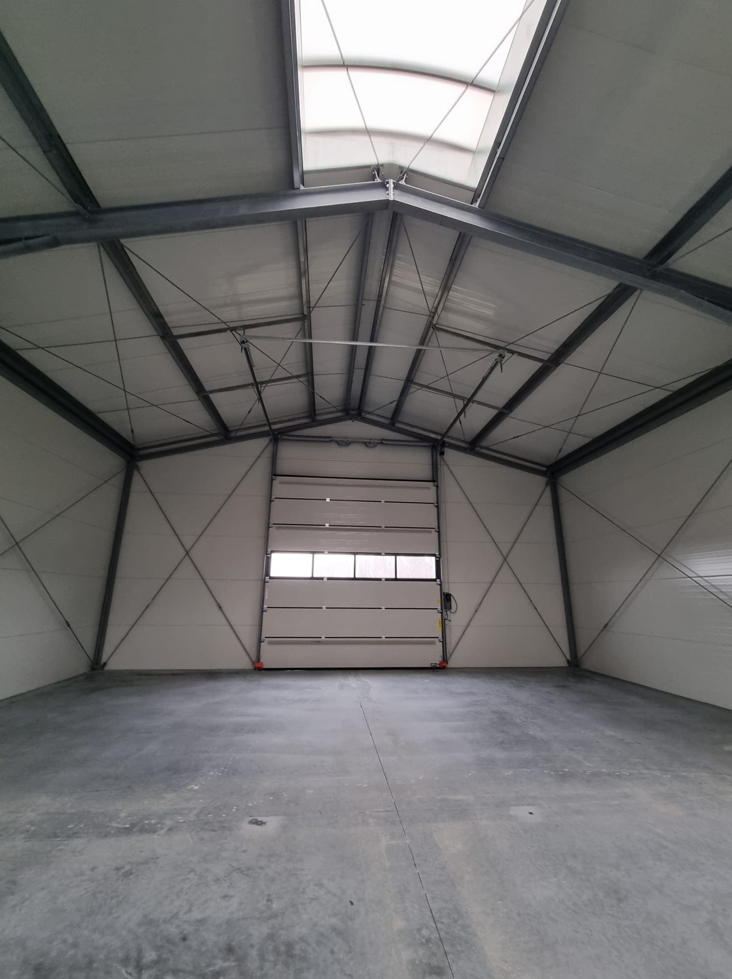 Wnętrze pustego, nowo wybudowanego magazynu z betonową posadzką, stalową konstrukcją nośną i częściowo otwartą bramą segmentową.