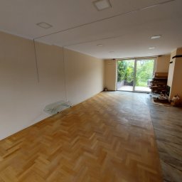 HORIZON VITALII VDOVYCHENKO - Salon z parkietem w jodełkę, jasne ściany, przesuwne drzwi balkonowe. Widoczne szklane półki i elementy instalacji pod sufitem. Częściowo widoczny kominek.