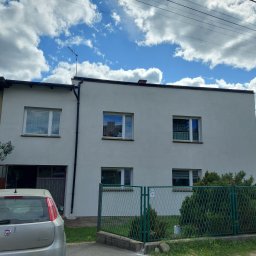 HORIZON VITALII VDOVYCHENKO - Dwukondygnacyjny, biały dom z oknami, zielonym ogrodzeniem i fragmentem samochodu. Błękitne niebo z obłokami w tle. Widoczne elementy elewacji i instalacji.