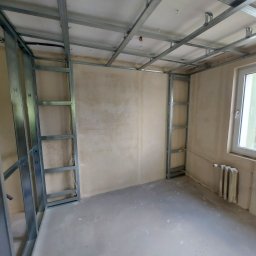 HORIZON VITALII VDOVYCHENKO - Montaż metalowej konstrukcji ścianki działowej i sufitu podwieszanego w surowym wnętrzu. Widoczne profile i płyta OSB, okno i grzejnik.