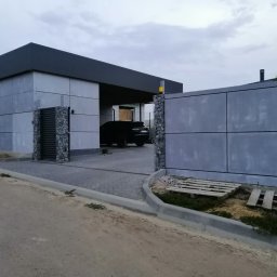 HORIZON VITALII VDOVYCHENKO - Nowoczesna elewacja budynku z szarych paneli i gabionów. Brama wjazdowa z elementami kamiennymi. Na podjeździe zaparkowany czarny samochód.