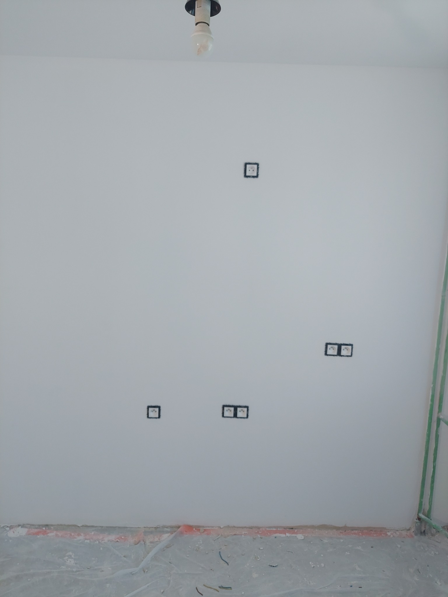 Biała ściana z gładzią i gniazdkami elektrycznymi. Podłoga zabezpieczona folią. Widoczna żarówka i fragment instalacji.