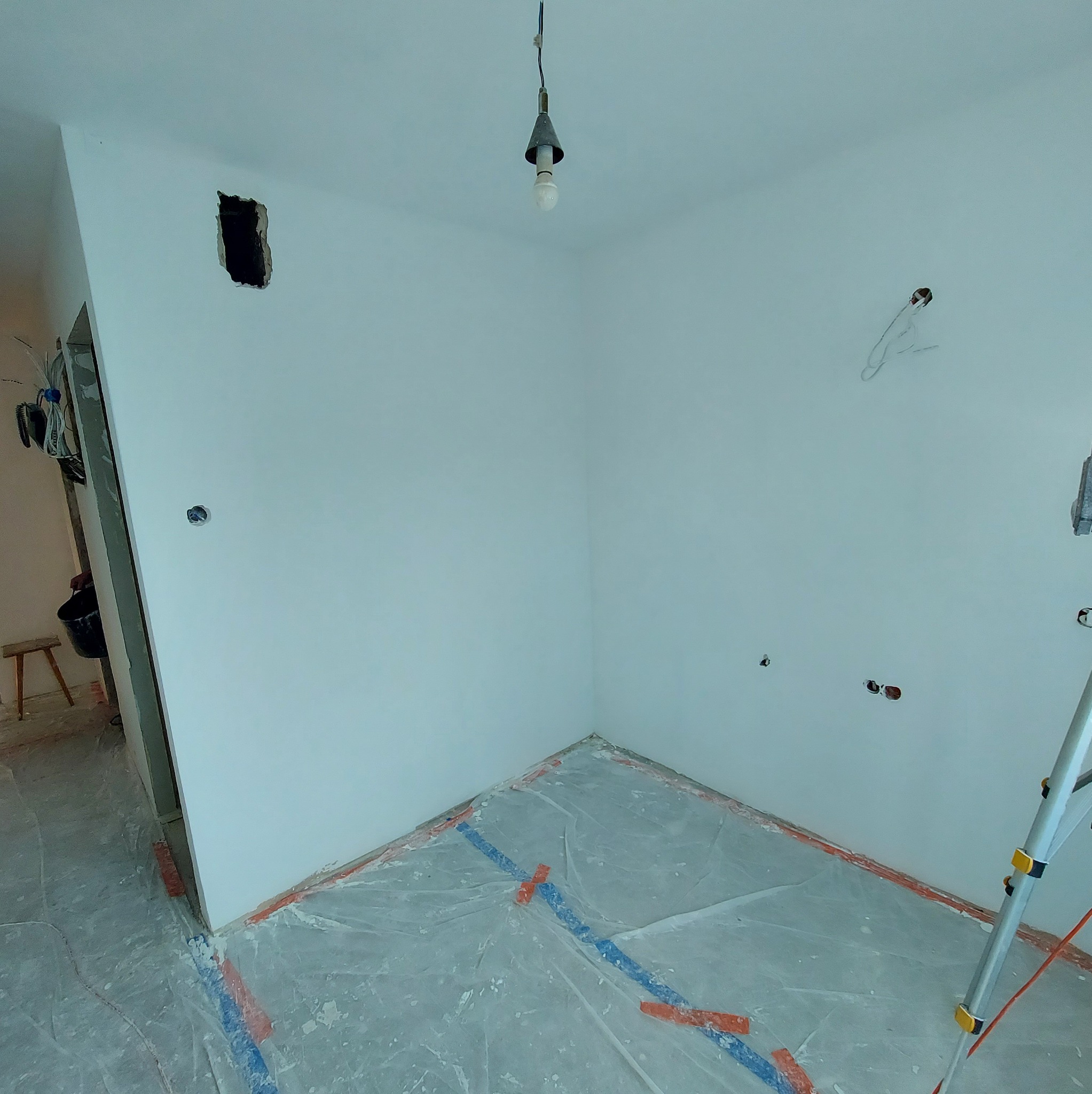 Wnętrze w trakcie remontu: białe ściany z widocznymi otworami na instalacje, zabezpieczona folią podłoga i drabina. Sufit z żarówką i wystającymi kablami.