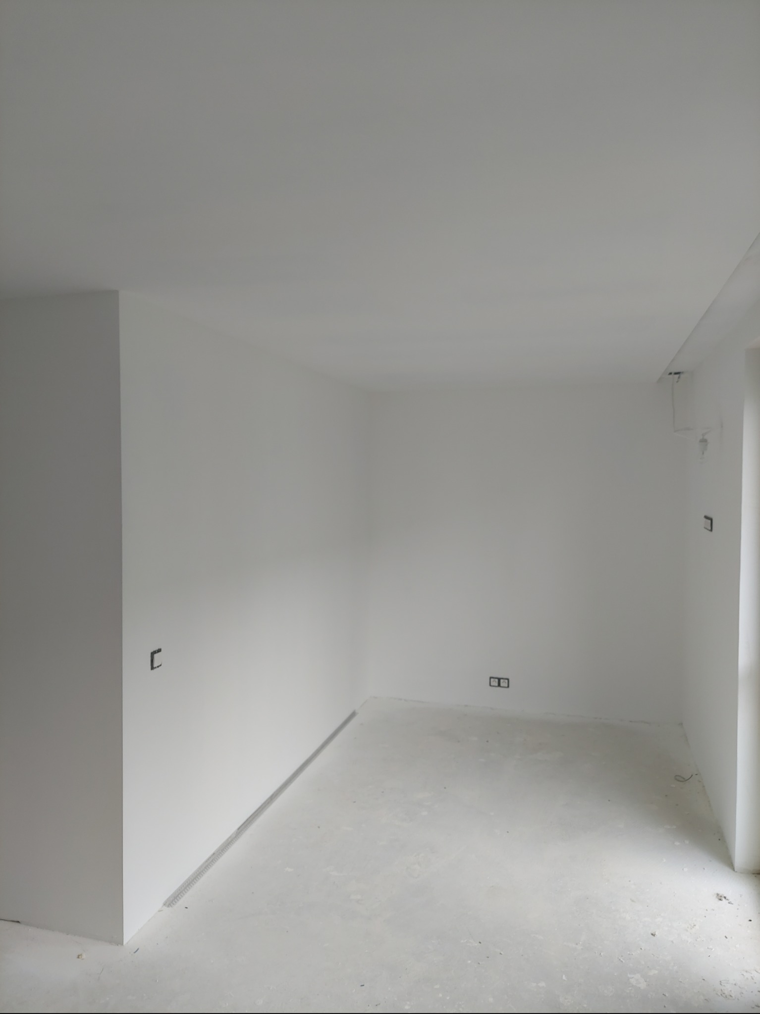 Białe, puste wnętrze po remoncie z gładkimi ścianami, gotowe do wykończenia. Widoczne puszki elektryczne i listwa podłogowa.