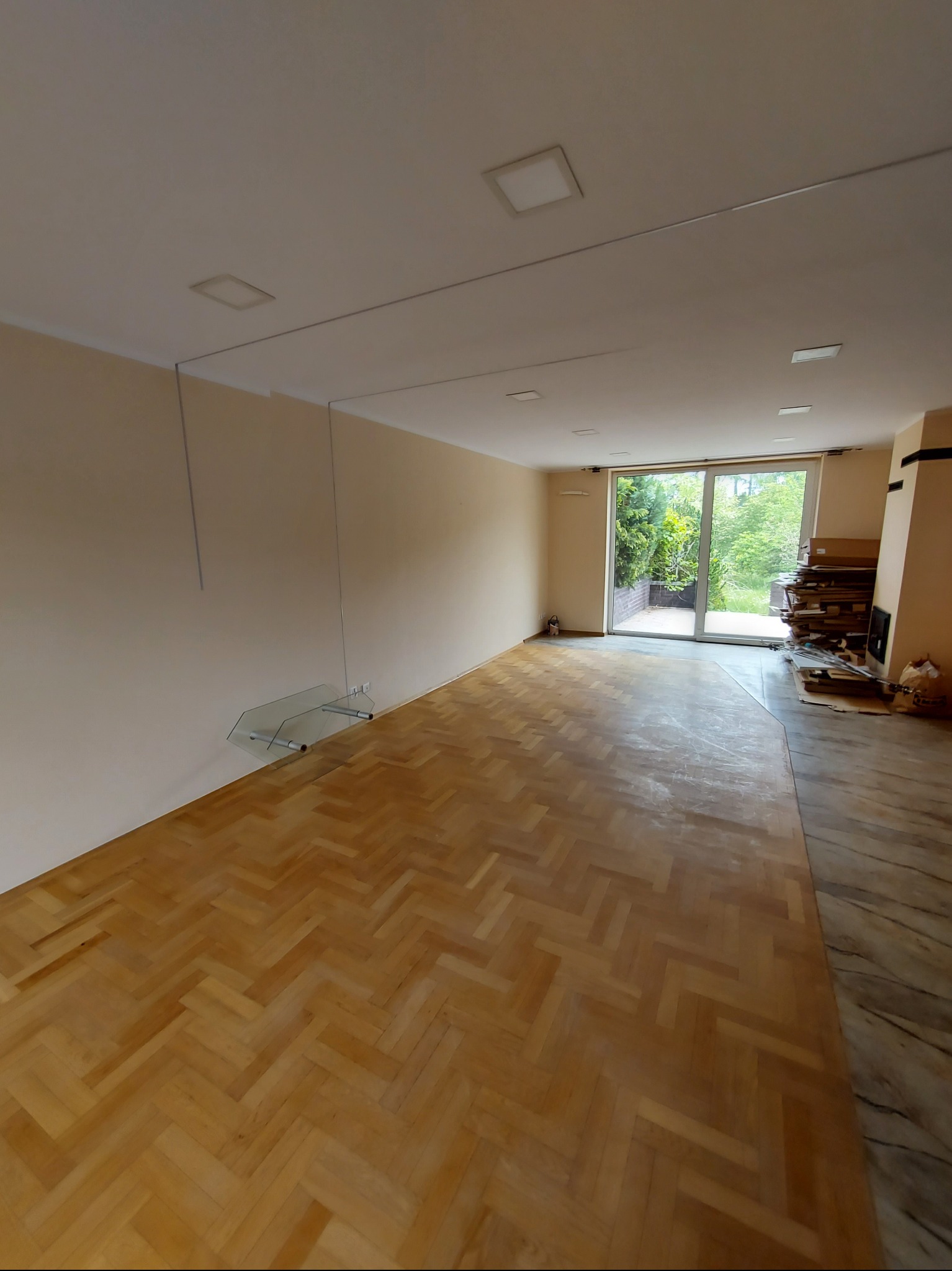 Salon z parkietem w jodełkę, jasne ściany, przesuwne drzwi balkonowe. Widoczne szklane półki i elementy instalacji pod sufitem. Częściowo widoczny kominek.