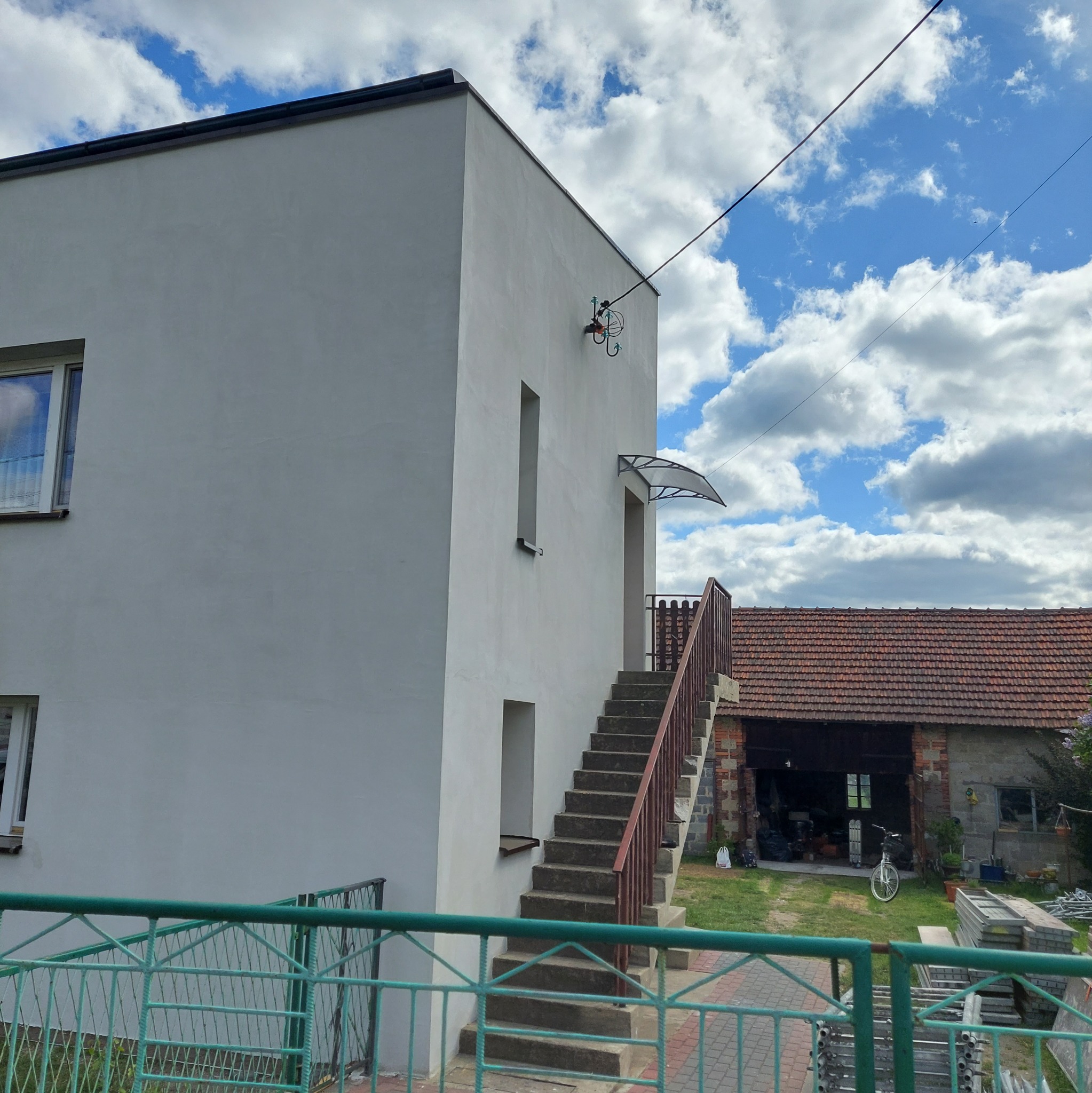 Biały budynek z zewnętrznymi schodami i zadaszeniem nad drzwiami, widok z dołu, błękitne niebo z chmurami w tle. Widoczne okna i elementy wykończeniowe.