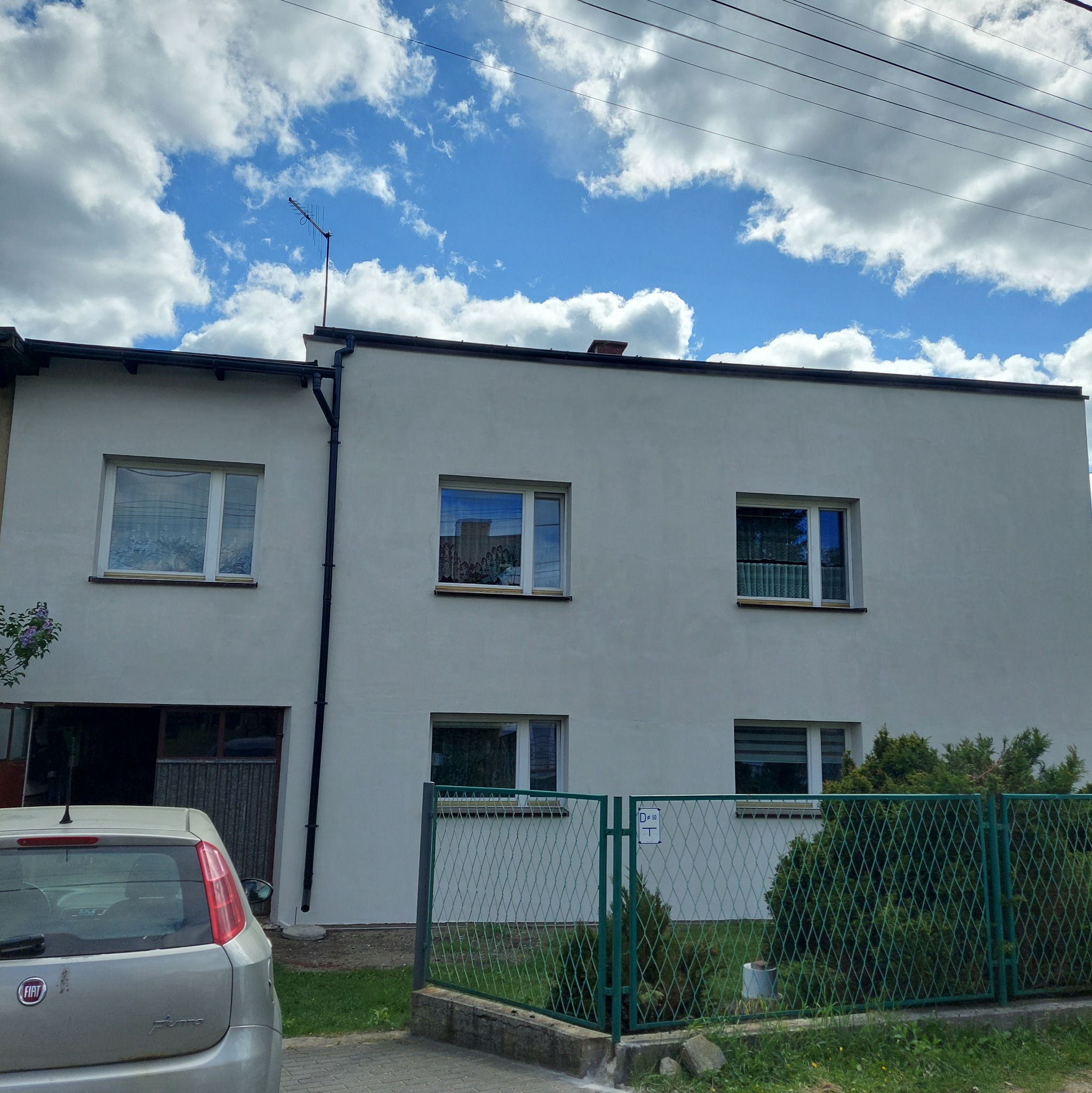 Dwukondygnacyjny, biały dom z oknami, zielonym ogrodzeniem i fragmentem samochodu. Błękitne niebo z obłokami w tle. Widoczne elementy elewacji i instalacji.